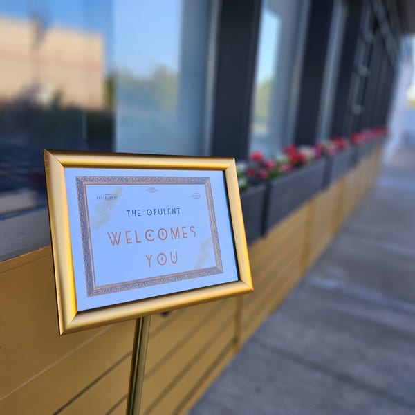 A golden framed sign outside The Opulent Restaurant and Lounge reads "The Opulent Welcomes You".