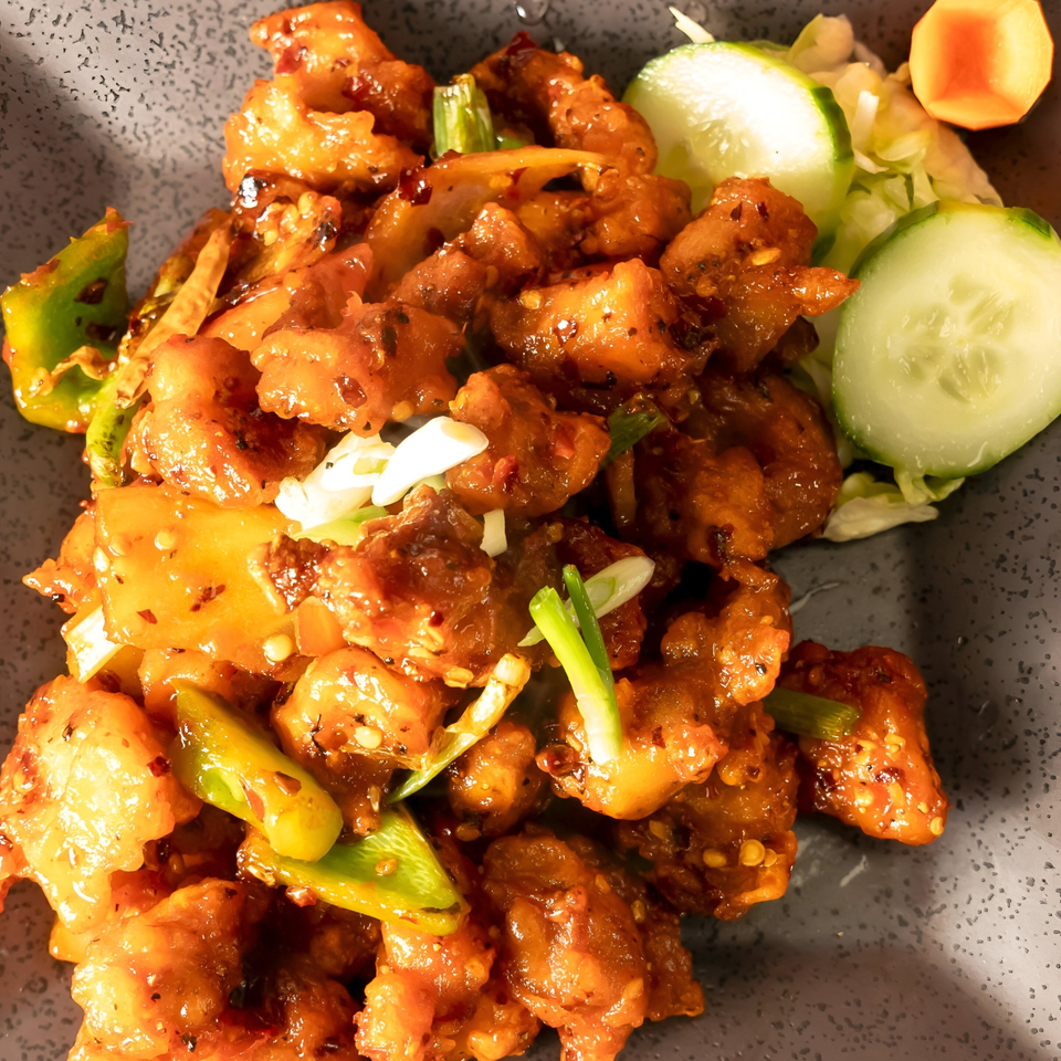 Close-up of Indian chili chicken with bell peppers, onions, and a side of cucumber slices.