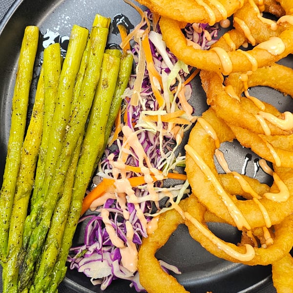 Grilled asparagus, colorful coleslaw, and crispy onion rings drizzled with sauce on a dark plate.